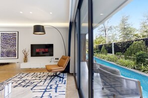 Ein modernes Wohnzimmer mit großem Fernseher, einem bequemen Sessel, Blick auf einen Pool und einem minimalistischen Design.