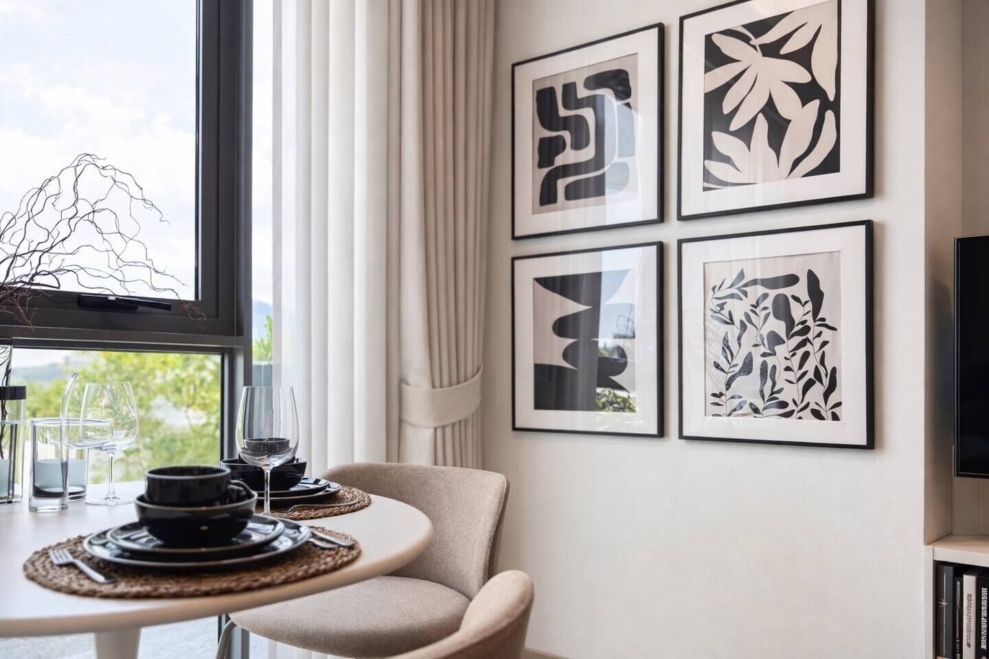 A dining area with a table set for two, framed artwork on the wall, and a view through the window.