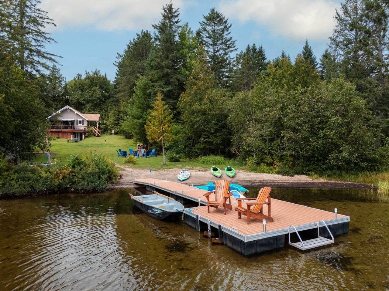 A wooden dock with chairs and kayaks, a house in the background, and a forested area.
