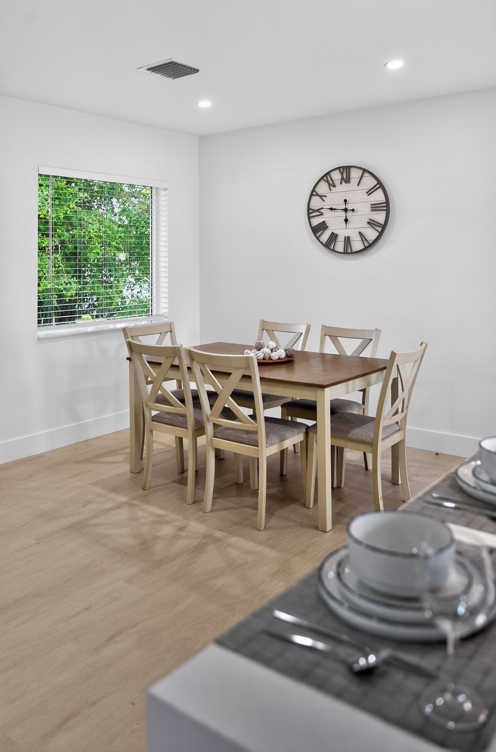 Een eetruimte met een houten tafel en stoelen, een wandklok en een raam met beplanting erbuiten.