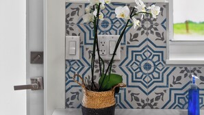 Una cucina con un piano di lavoro bianco, un poster di un mercato di frutta appeso al muro, uno schienale decorato e una pianta di orchidea in vaso.