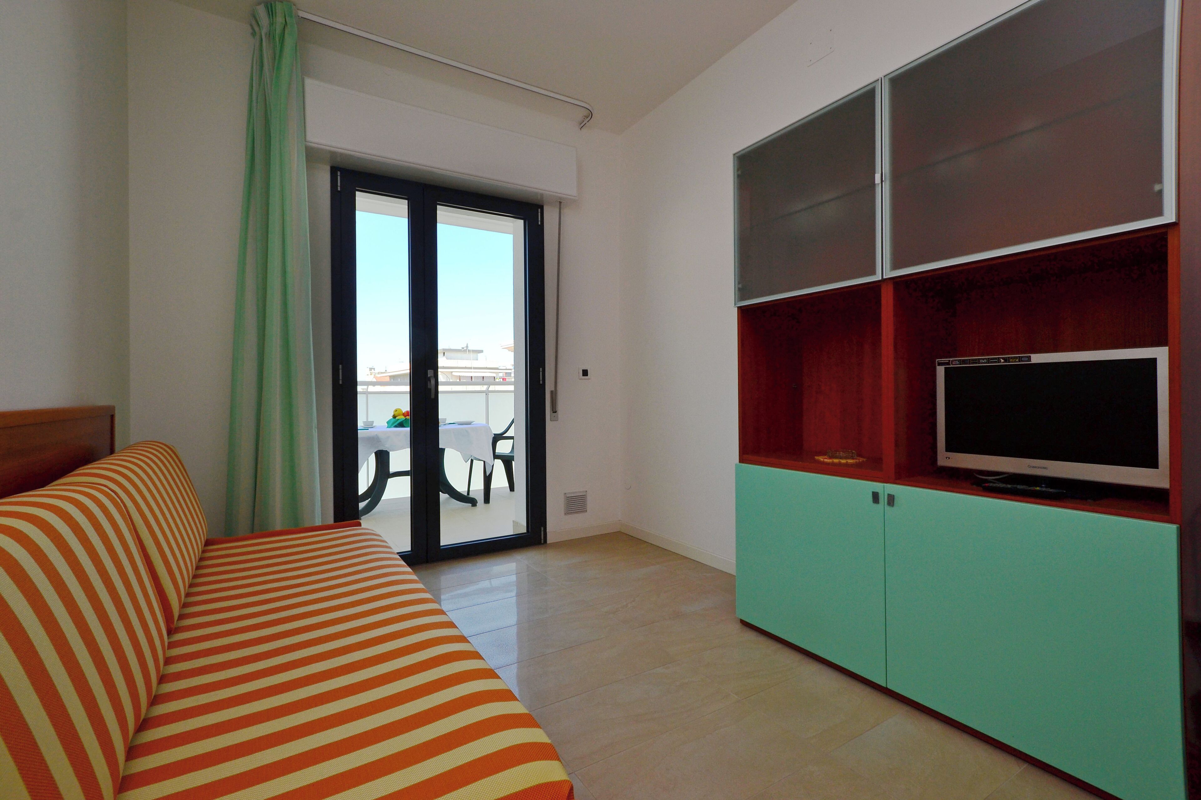 A hotel room with a bed, a television on a cabinet, a sliding glass door to the balcony, and a dining table outside.