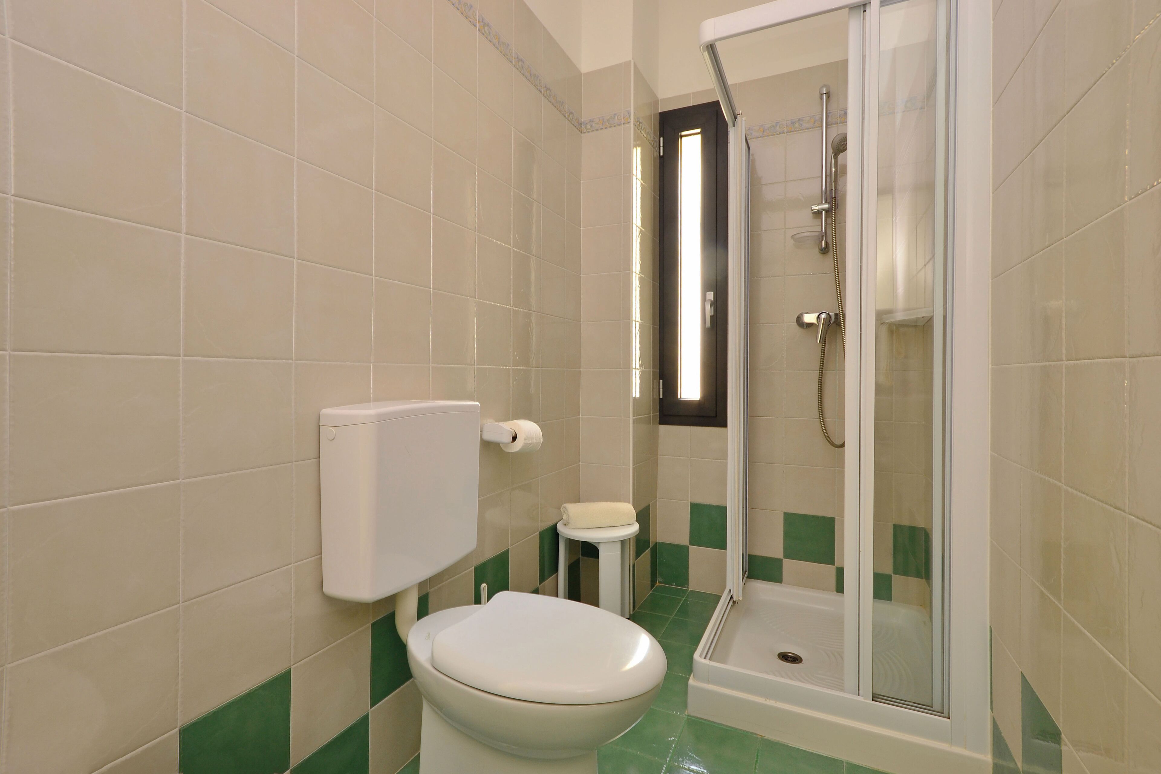 Une salle de bain comprenant des toilettes, un petit tabouret, une douche fermée par une paroi de verre et un porte-rouleau de papier toilette.