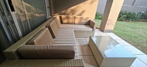 Un área de estar al aire libre con luz solar, que incluye un sofá y una mesa de vidrio, rodeada por una estructura de concreto y un césped.