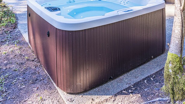 A hot tub in a forested area with a wooden deck, outdoor seating, and a barbecue grill.