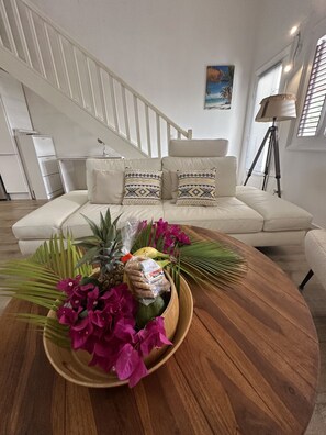 Ein Wohnzimmer mit einem weißen Sofa, einem Couchtisch mit Obst und Blumen, und einer Treppe im Hintergrund.