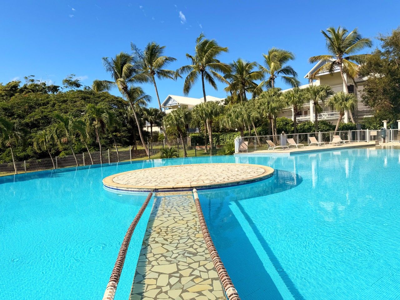 Une piscine d’un bleu éclatant, bordée d’une terrasse carrelée, entourée de palmiers et avec un bâtiment visible au loin.