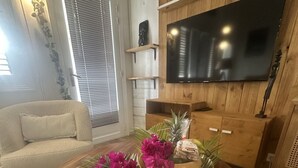 Olohuoneessa on pyöreä puinen sohvapöytä, beige sohva, seinälle kiinnitetty litteänäyttöinen televisio ja ikkuna, jossa on sälekaihtimet.