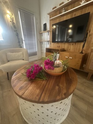 Ein Wohnzimmer mit rundem Holztisch, einem beigefarbenen Sofa, einem an einer Holzwand montierten Flachbildfernseher und einem Fenster mit Jalousien.