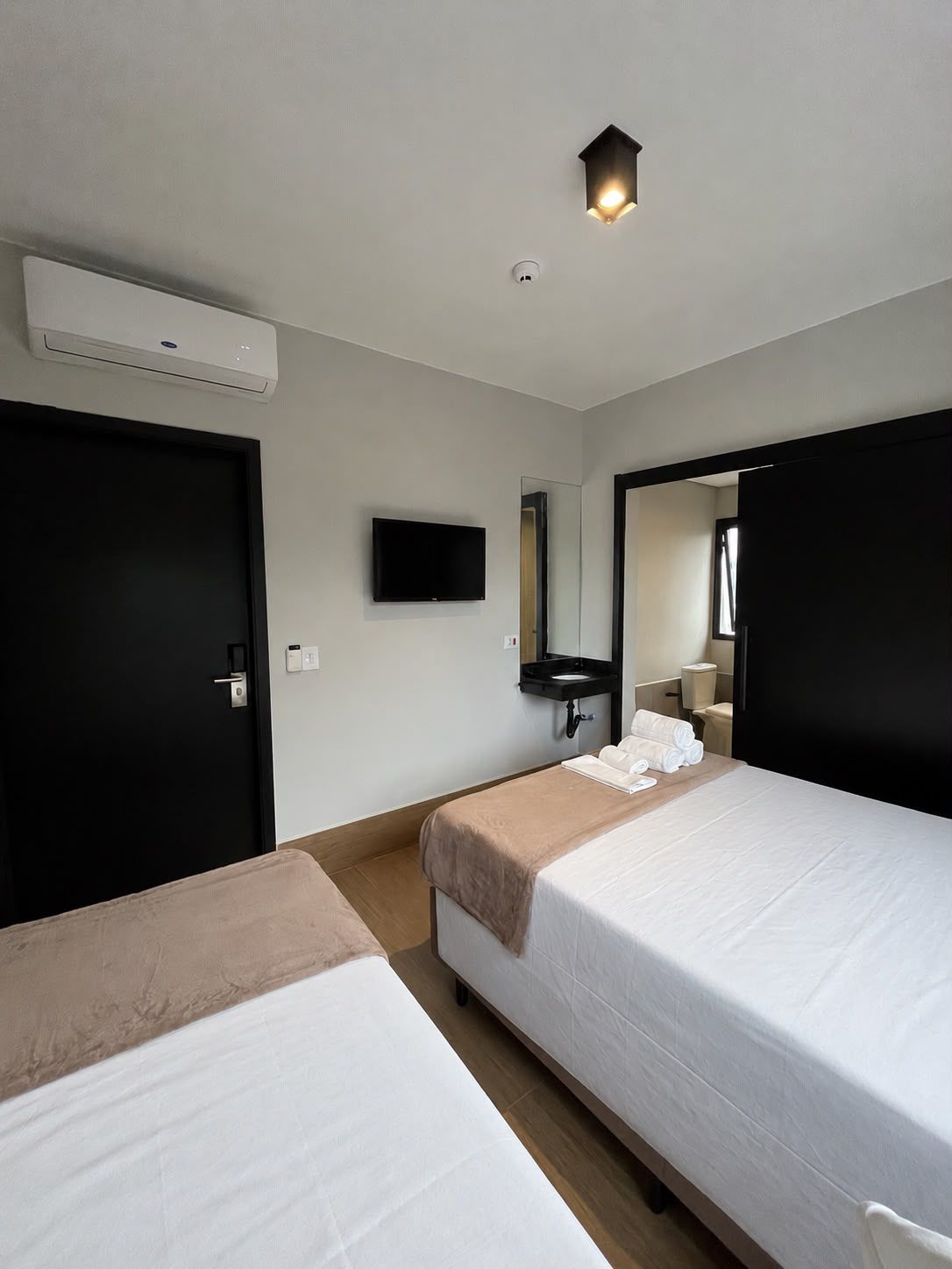 A hotel room with two beds, a flat-screen TV, a mirror, a bedside table with towels, and an air conditioning unit.