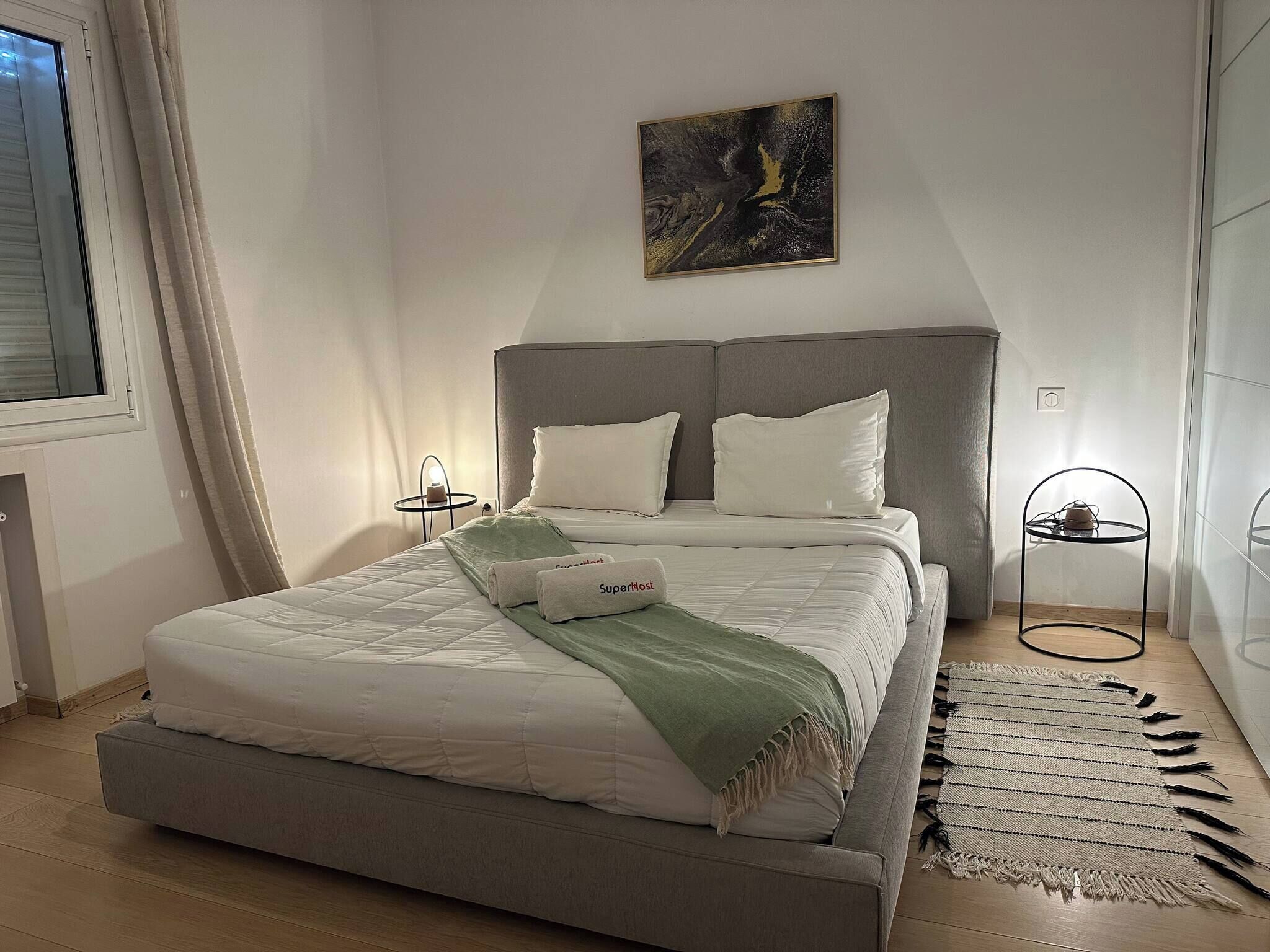 A bedroom with a bed, a bedside table, a lamp, a painting, and a window.