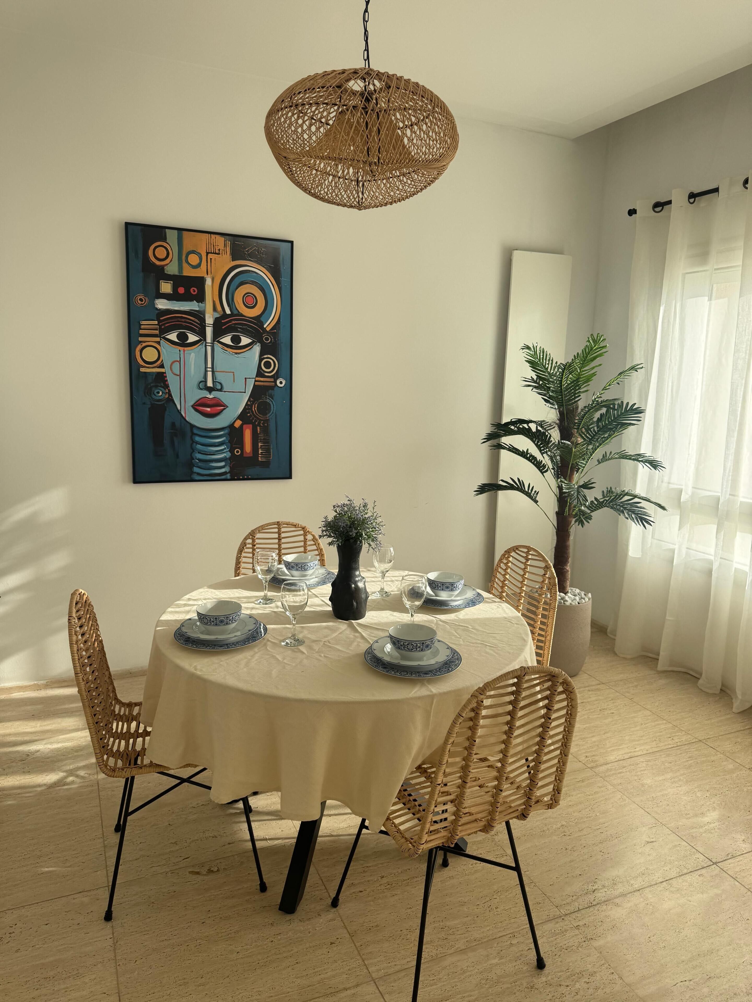 Un coin repas avec une table ronde dressée pour quatre personnes, entourée de chaises en osier, d’un luminaire suspendu en rotin, d’un grand tableau abstrait et d’une plante en pot.