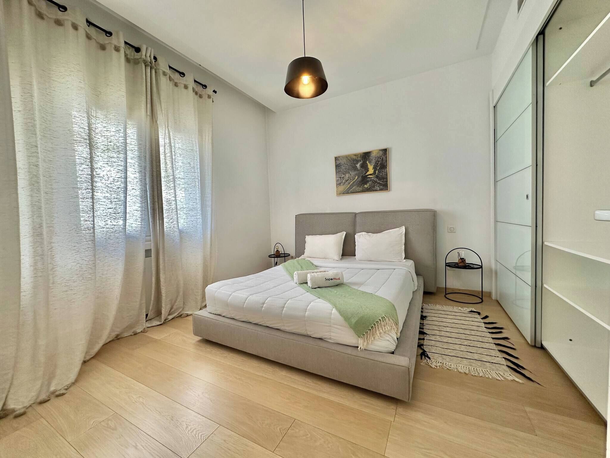 A modern bedroom with a bed, curtains, a painting, and a hanging light fixture.