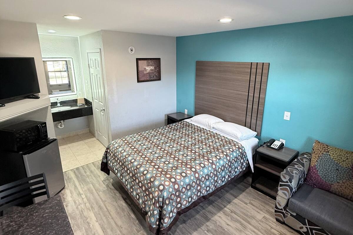 A hotel room with a bed, bedside table, TV, kitchenette, and a bathroom.