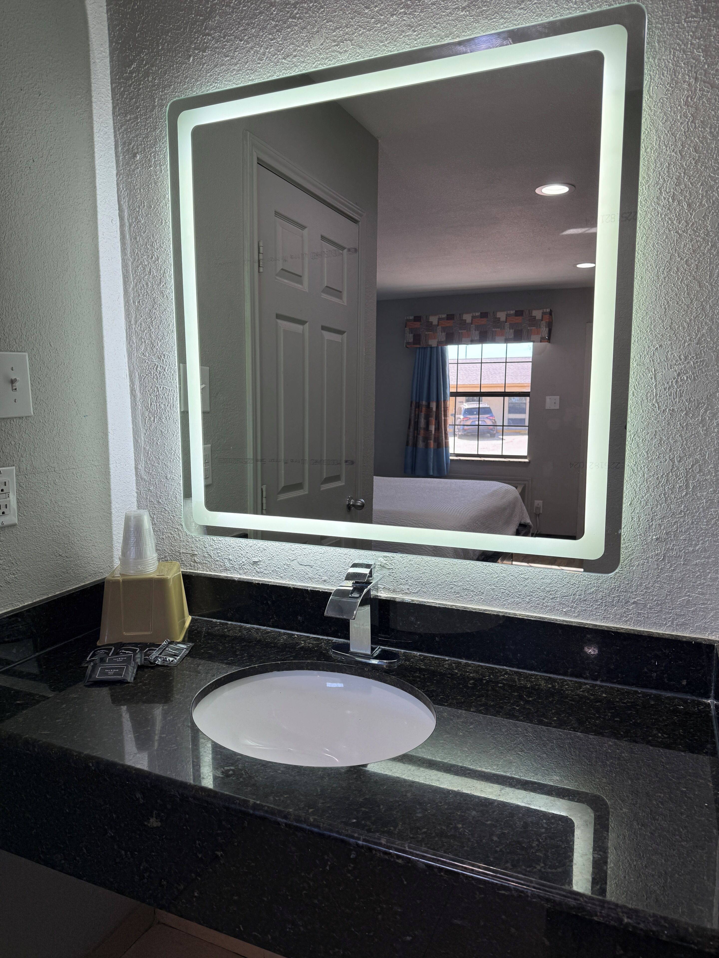 Baño de hotel con un espejo grande, un lavamanos, dispensador de toallas y una ventana con vistas al exterior.
