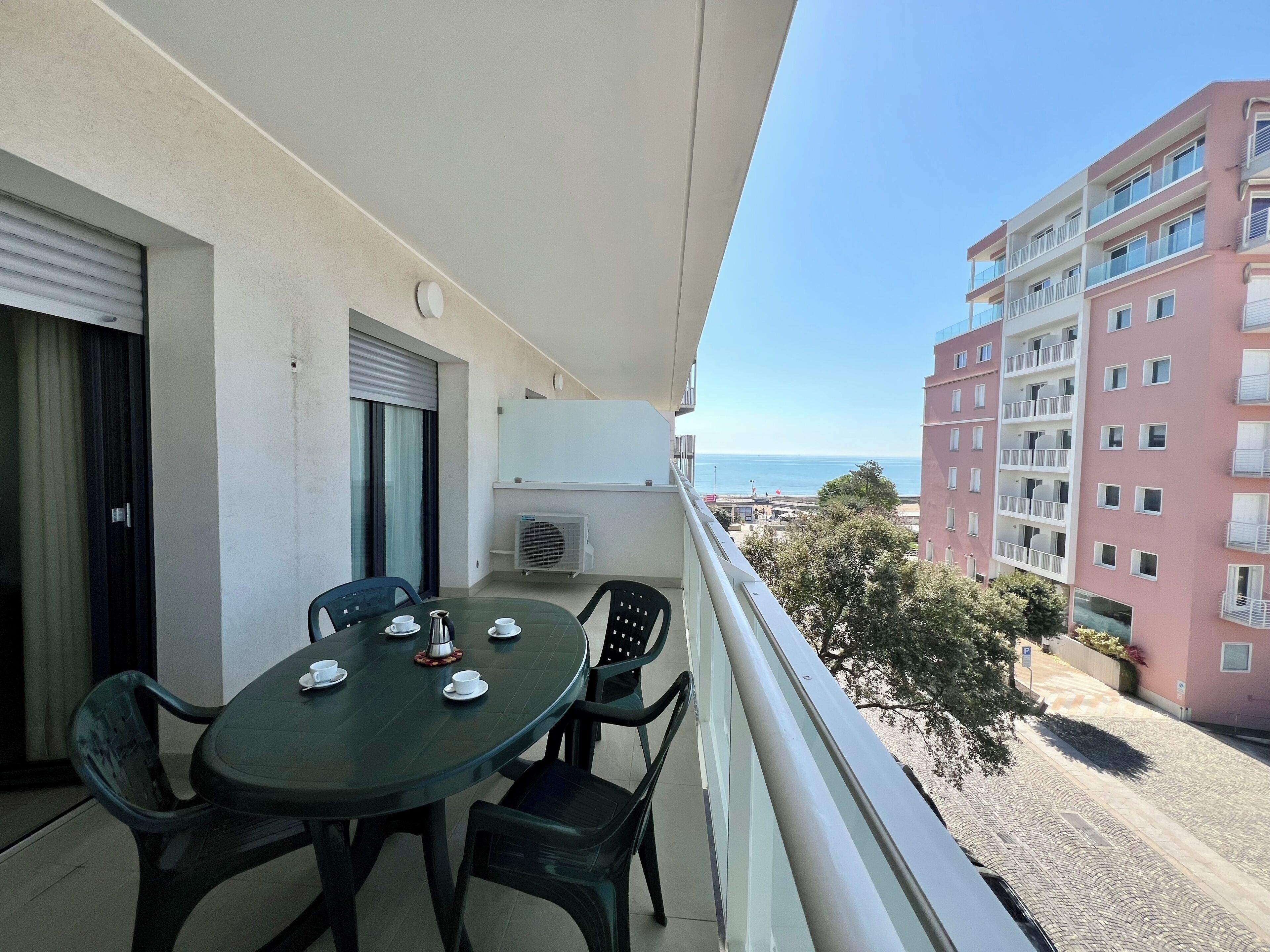 Een balkon met een tafel en stoelen, een airconditioning en uitzicht op de zee en gebouwen.