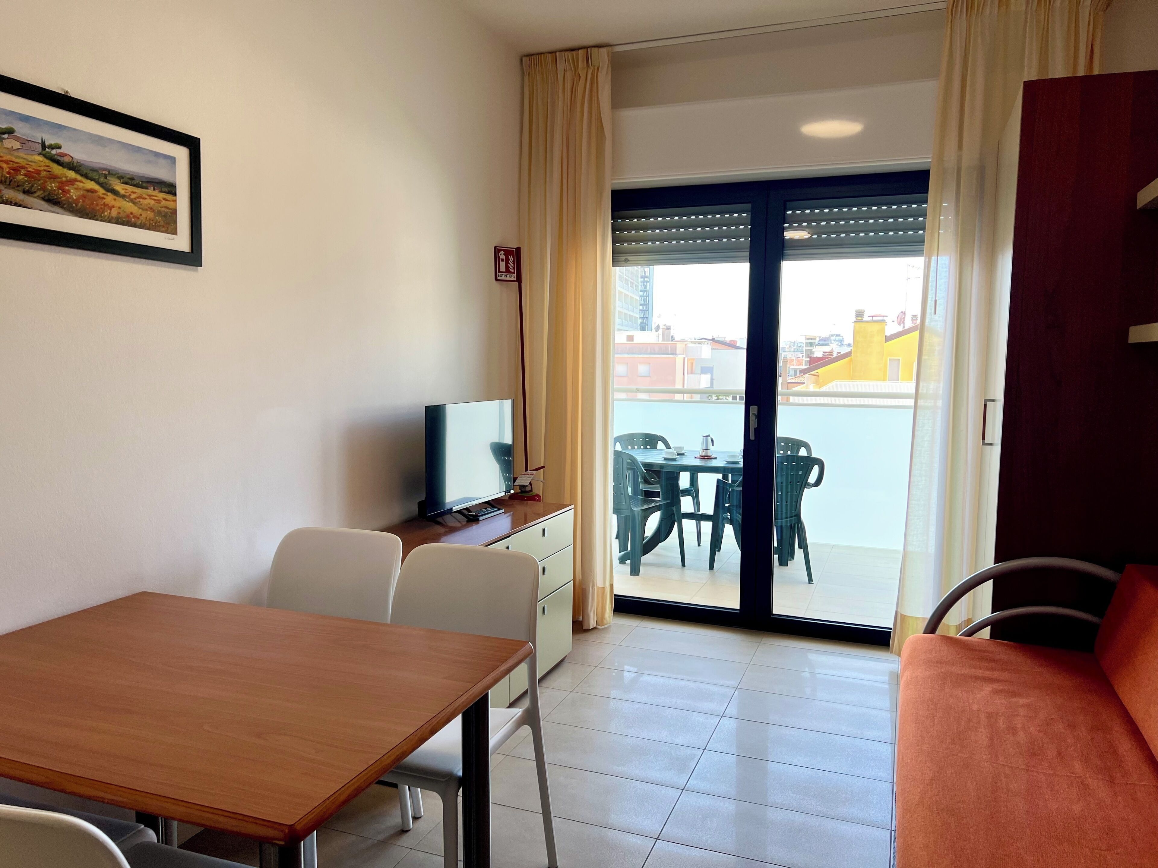 A hotel room with a wooden table, a TV, a balcony with a table and chairs, and a painting on the wall.