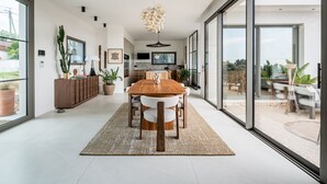 Un comedor moderno con mesa y sillas de madera, una lámpara de araña grande y vistas a un patio exterior a través de puertas de cristal amplias.