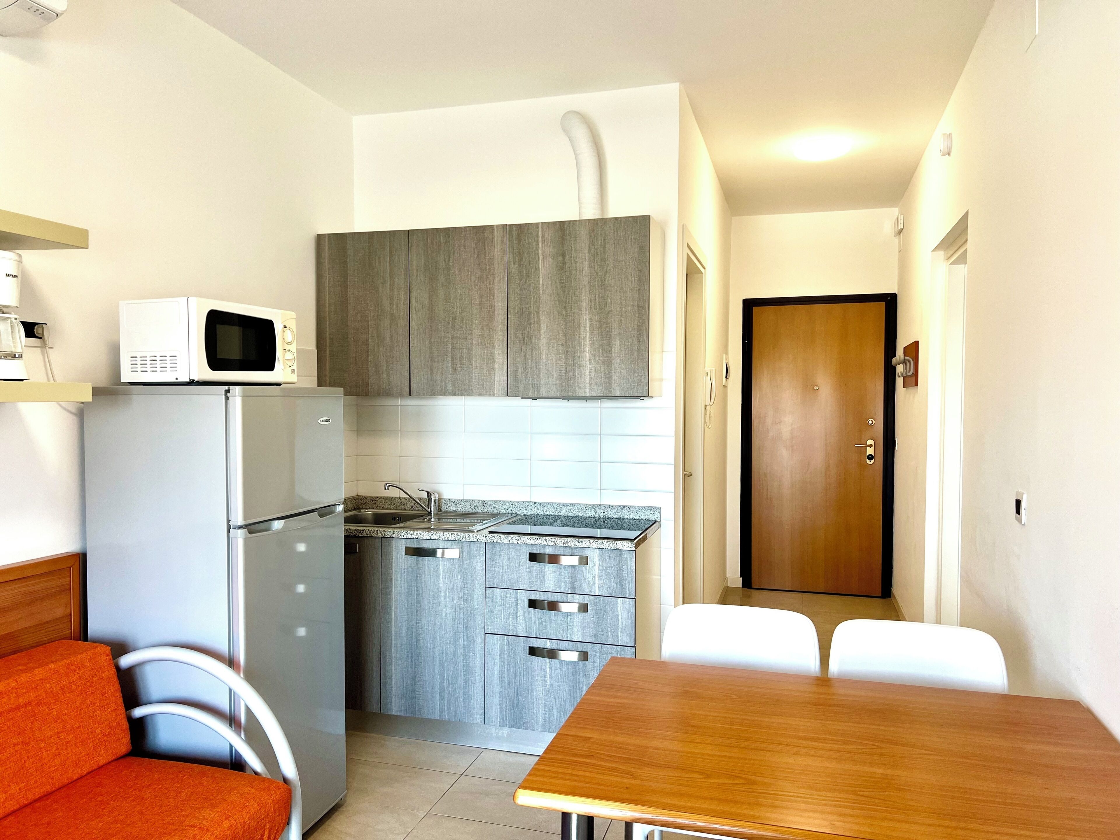 A compact kitchen with a microwave, refrigerator, and a small sink. There is a wooden dining table and two white chairs.