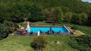 Ein rechteckiger Swimmingpool mit Liegestühlen und Sonnenschirmen, umgeben von einer Rasenfläche und Bäumen, mit einer Hügelkette im Hintergrund.