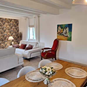 Ein Wohnzimmer mit einer Couch, einem roten Sessel, einem Holztisch mit Platzdeckchen, einem Bild an der Wand und einem Fenster mit Vorhängen.