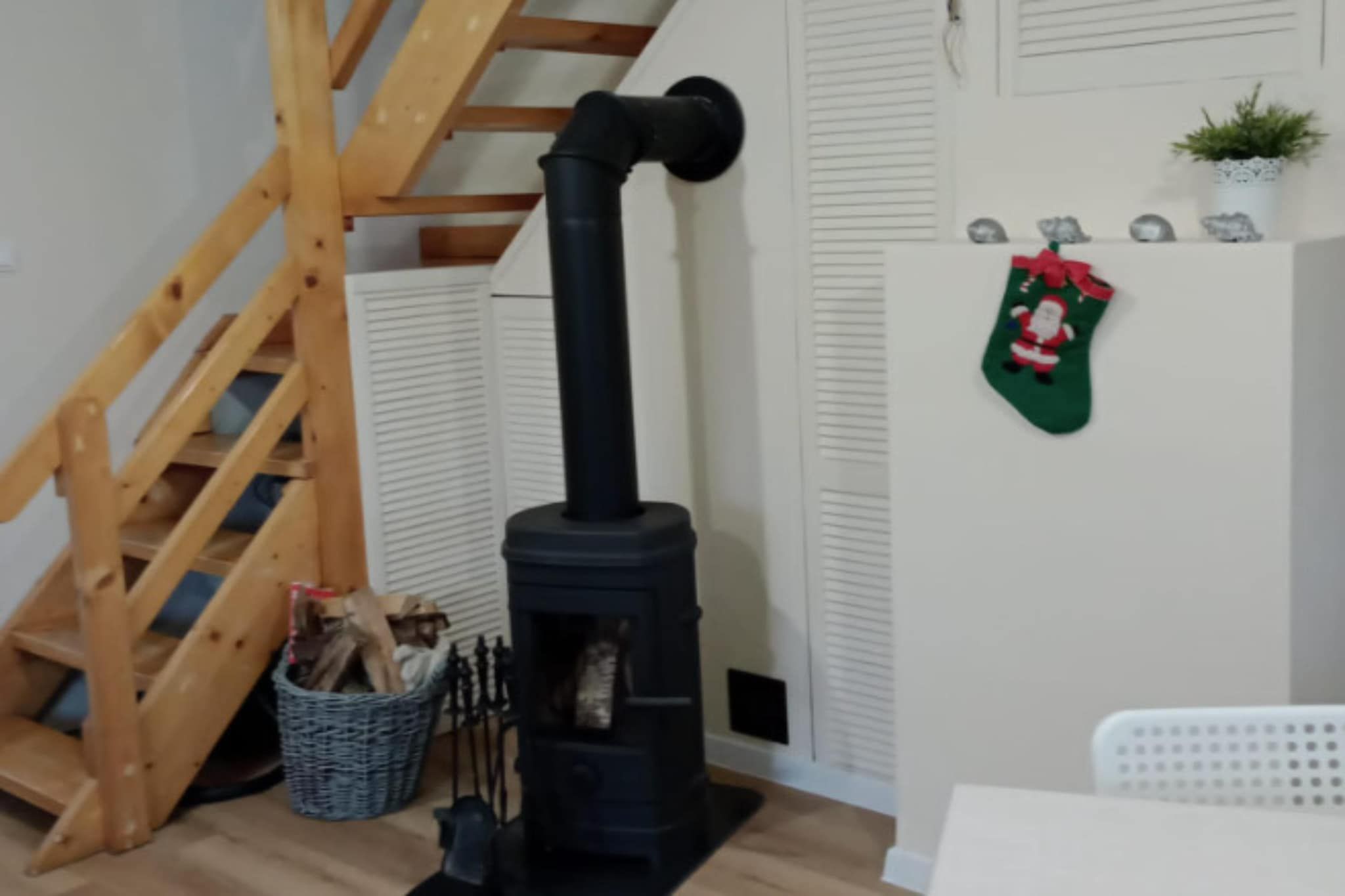A room with a black wood-burning stove, a wooden staircase, a basket of firewood, and a wall-mounted plant.