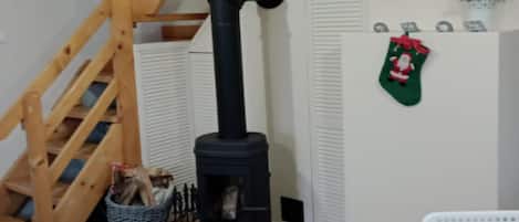 A room with a black wood-burning stove, a wooden staircase, a basket of firewood, and a wall-mounted plant.