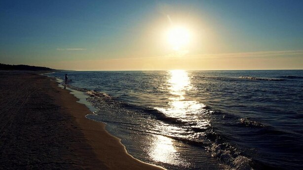 Ein Strand mit einer Person, die am Ufer entlanggeht, der Sonne, die sich im Wasser spiegelt, und einem klaren Himmel.