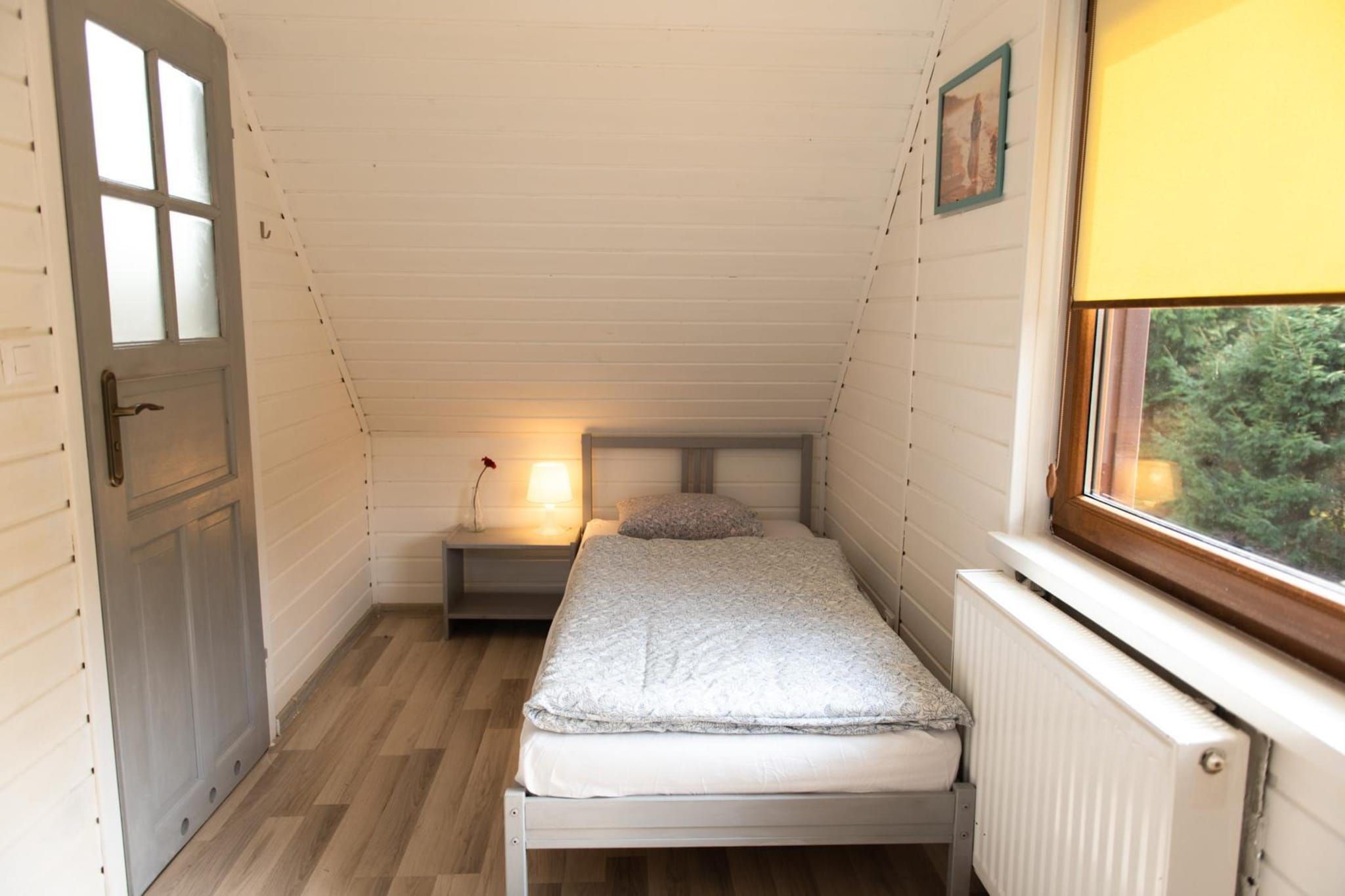 A narrow attic bedroom with a single bed, a nightstand, a lamp, and a window with a view of greenery.