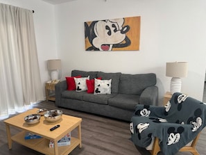 Un salón con sofá gris, almohadas rojas y blancas, una mesa de centro con cuencos y un control remoto, una lámpara y un cuadro en la pared.