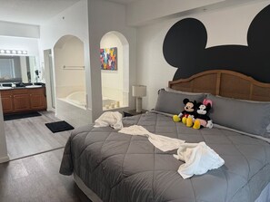 Un dormitorio con una cama, peluches de Mickey y Minnie Mouse, un cabecero, una mesita de noche, una lámpara, un baño con bañera y un tocador con espejo e iluminación.