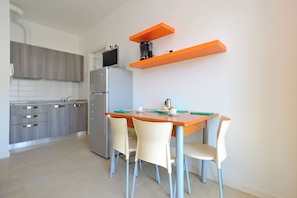 A modern kitchen with a dining table, chairs, a refrigerator, and orange floating shelves. - Comfortable Vacation Retreat Near the Beach (Lignano Sabbiadoro)