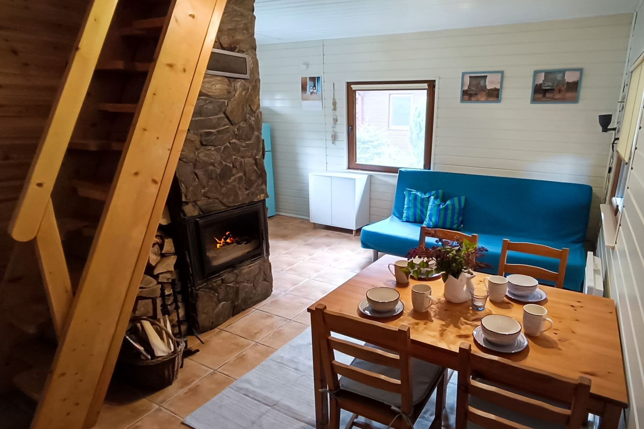 Une pièce chaleureuse avec une cheminée en pierre, une table à manger en bois dressée pour quatre personnes et un canapé bleu en arrière-plan.