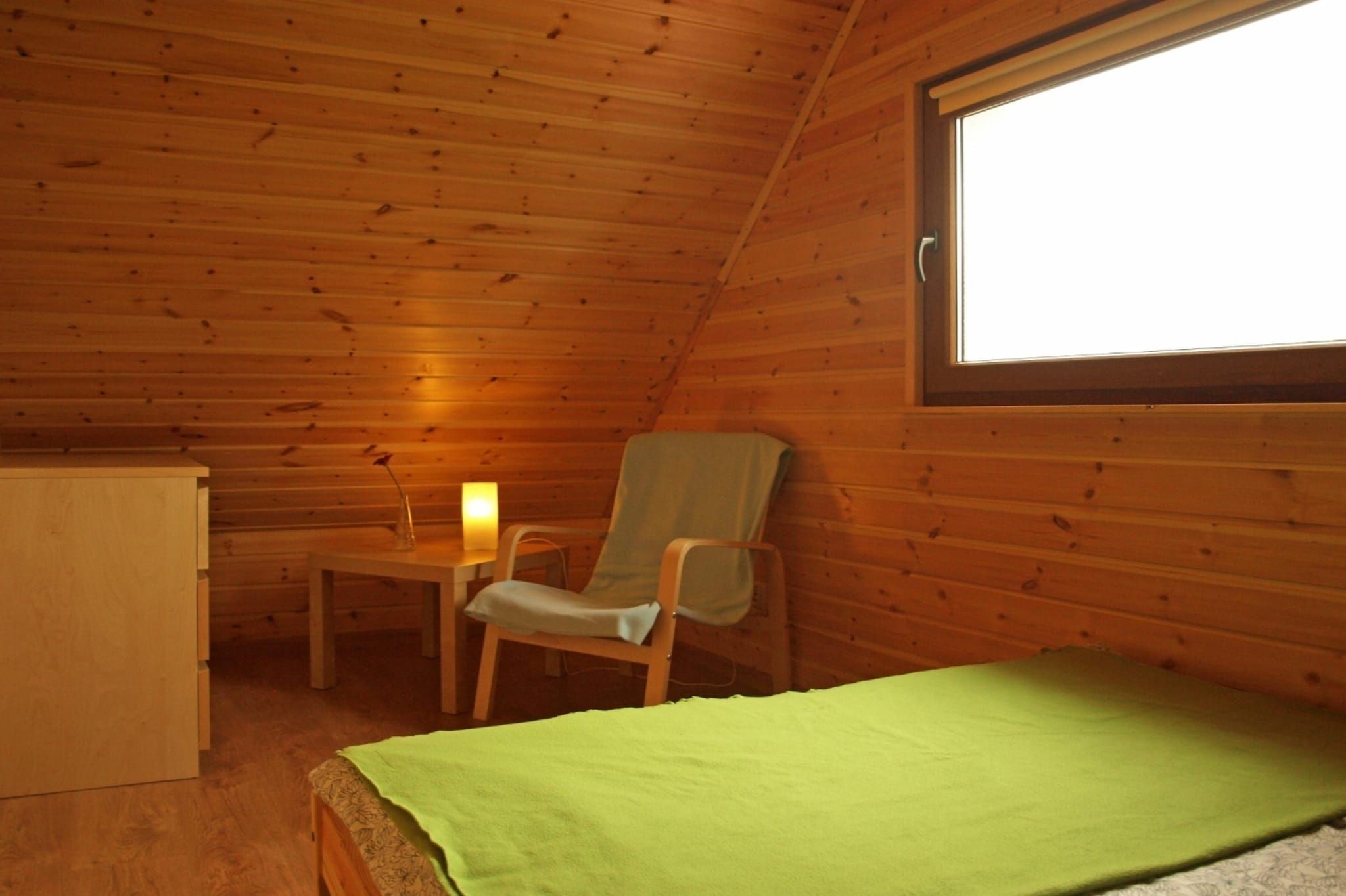 A small, single-room cabin with a wooden interior, a bed with a green cover, a chair, a table, and a window.