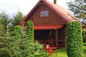A wooden cabin with a red roof, a covered porch, and a small table with a chair. - Holiday home with fireplace and large terrace (Kopalino)