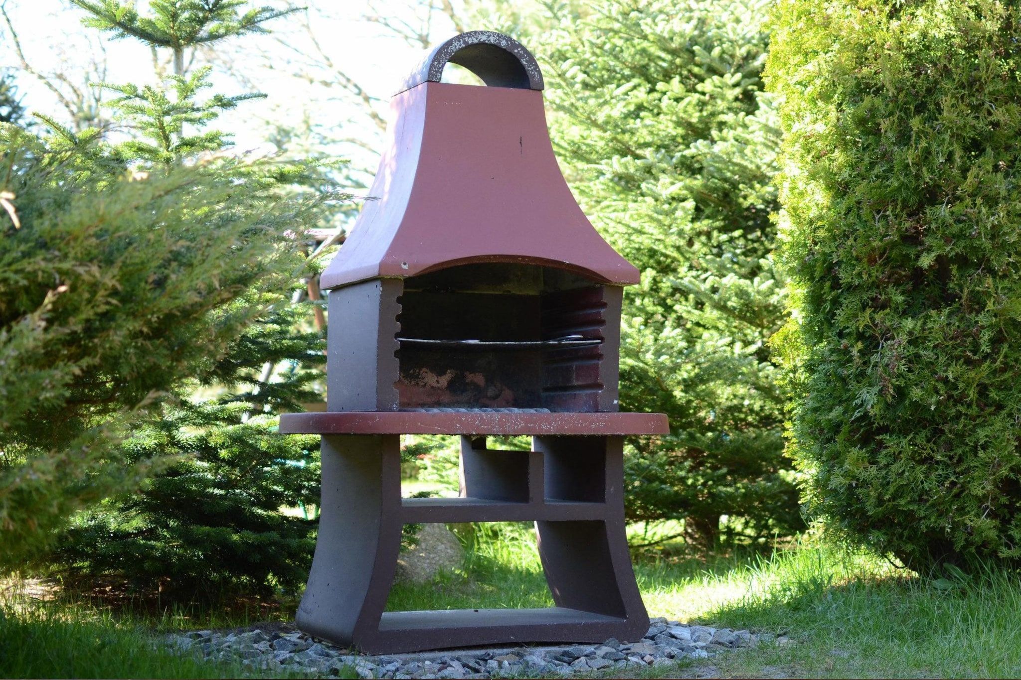 A barbecue grill with a chimney, set on a gravel base, surrounded by greenery.