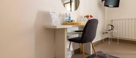 A room with a desk, chair, mirror, flat-screen TV, radiator, and a small table with a kettle.