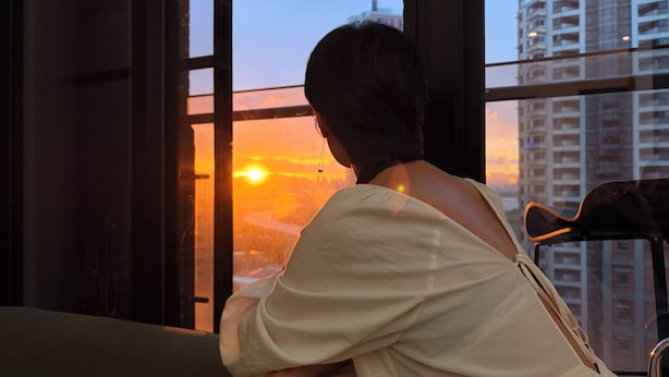 A person sitting by a window with a view of a sunset and high-rise buildings. - Luxury Sunset River View / Quiet & Convenient / Ximen / Monthly Rentals (Taipei City)