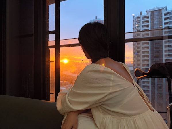 Une personne assise près d’une fenêtre, avec vue sur un coucher de soleil et des immeubles de grande hauteur.