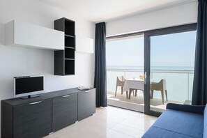 A modern room with a flat-screen TV on a dark cabinet, a balcony with a table and chairs, and a view of the sea. - Well-equipped Flat Just 50m From the Beach (Lignano Sabbiadoro)