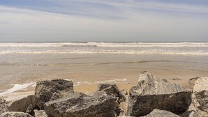 Ein Strand mit Wellen, blauem Himmel und großen Felsen im Vordergrund.
