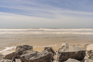 Ein Strand mit Wellen, blauem Himmel und großen Felsen im Vordergrund.