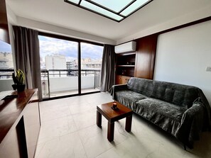 A modern living room with a sofa, a small table, a potted plant, and a large window with curtains. - STAY Finikoudes Nest 1 (Larnaca)