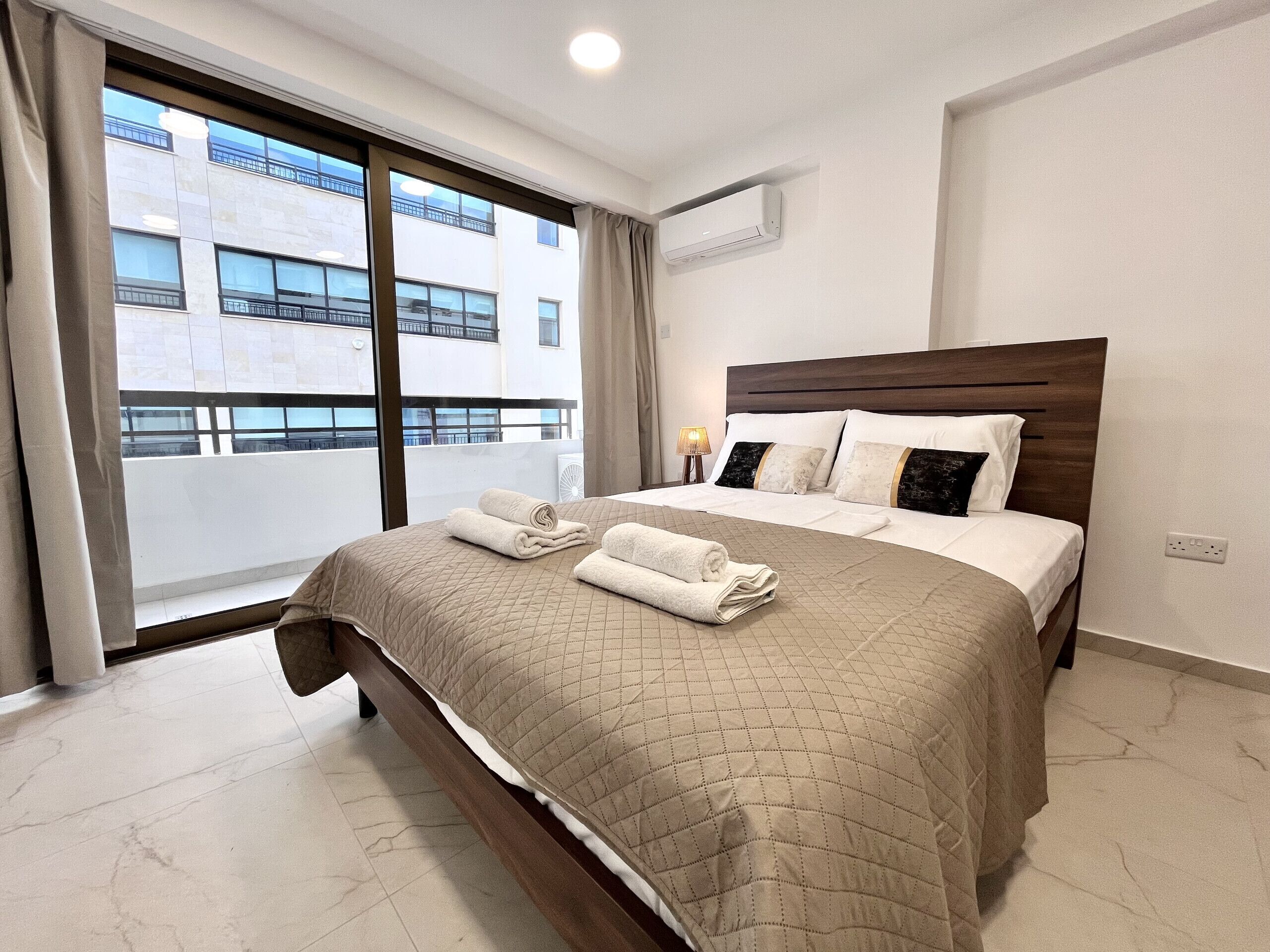 Quarto de hotel com uma cama grande, janela com vista para edifícios, ar condicionado e abajur.