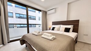 A hotel room with a large bed, a window with a view of buildings, an air conditioning unit, and a bedside lamp.
