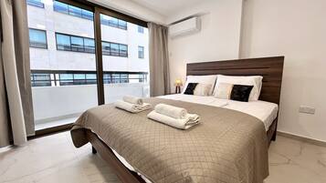 Quarto de hotel com uma cama grande, janela com vista para edifícios, ar condicionado e abajur.