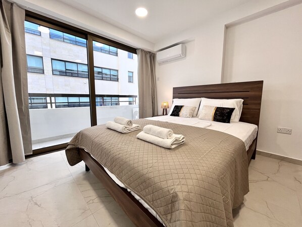Quarto de hotel com uma cama grande, janela com vista para edifícios, ar condicionado e abajur.