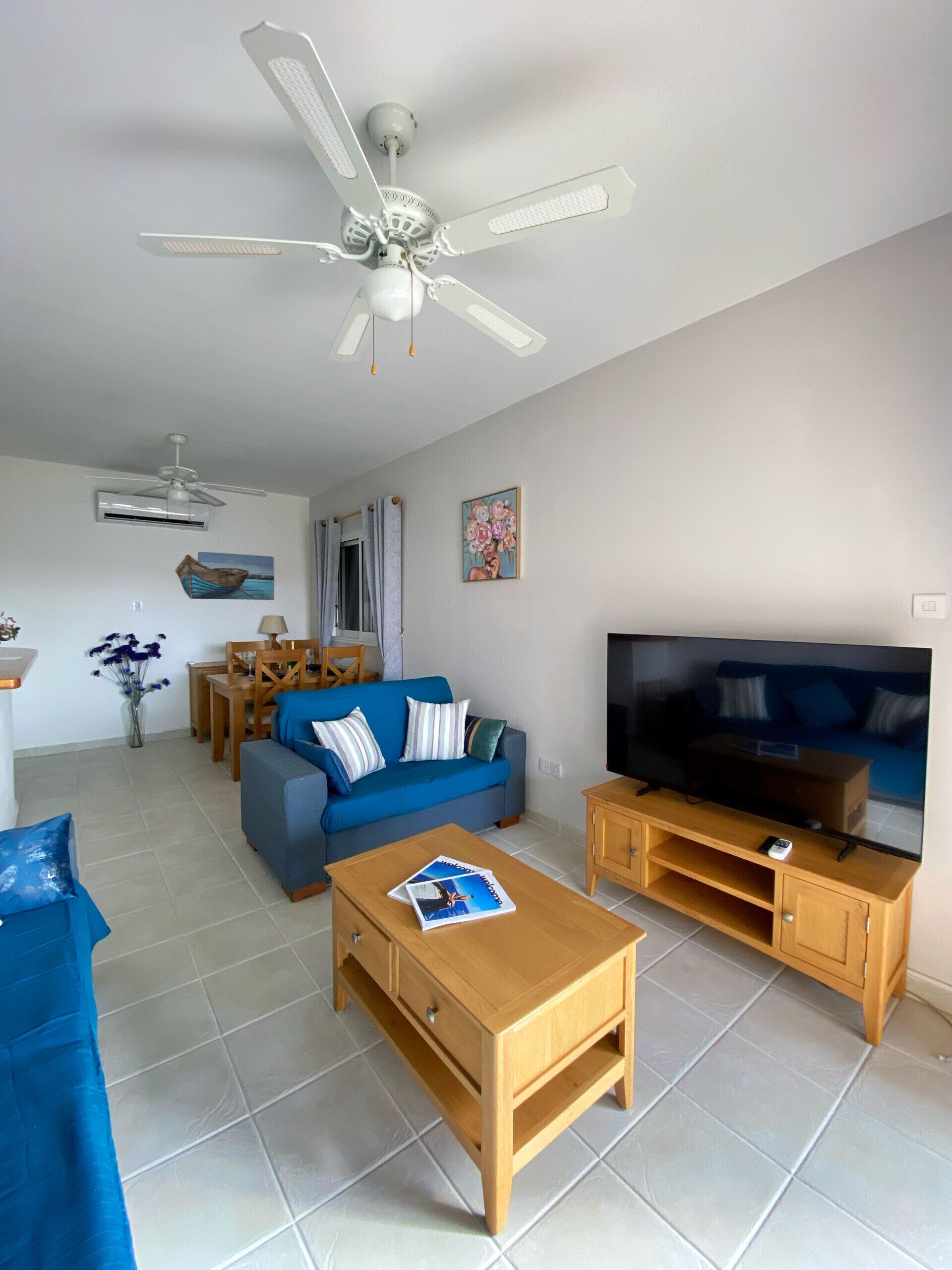 Ein Wohnzimmer mit Sofa, Couchtisch, TV-Möbel, Deckenventilator, Essbereich und Kunstwerken an den Wänden.
