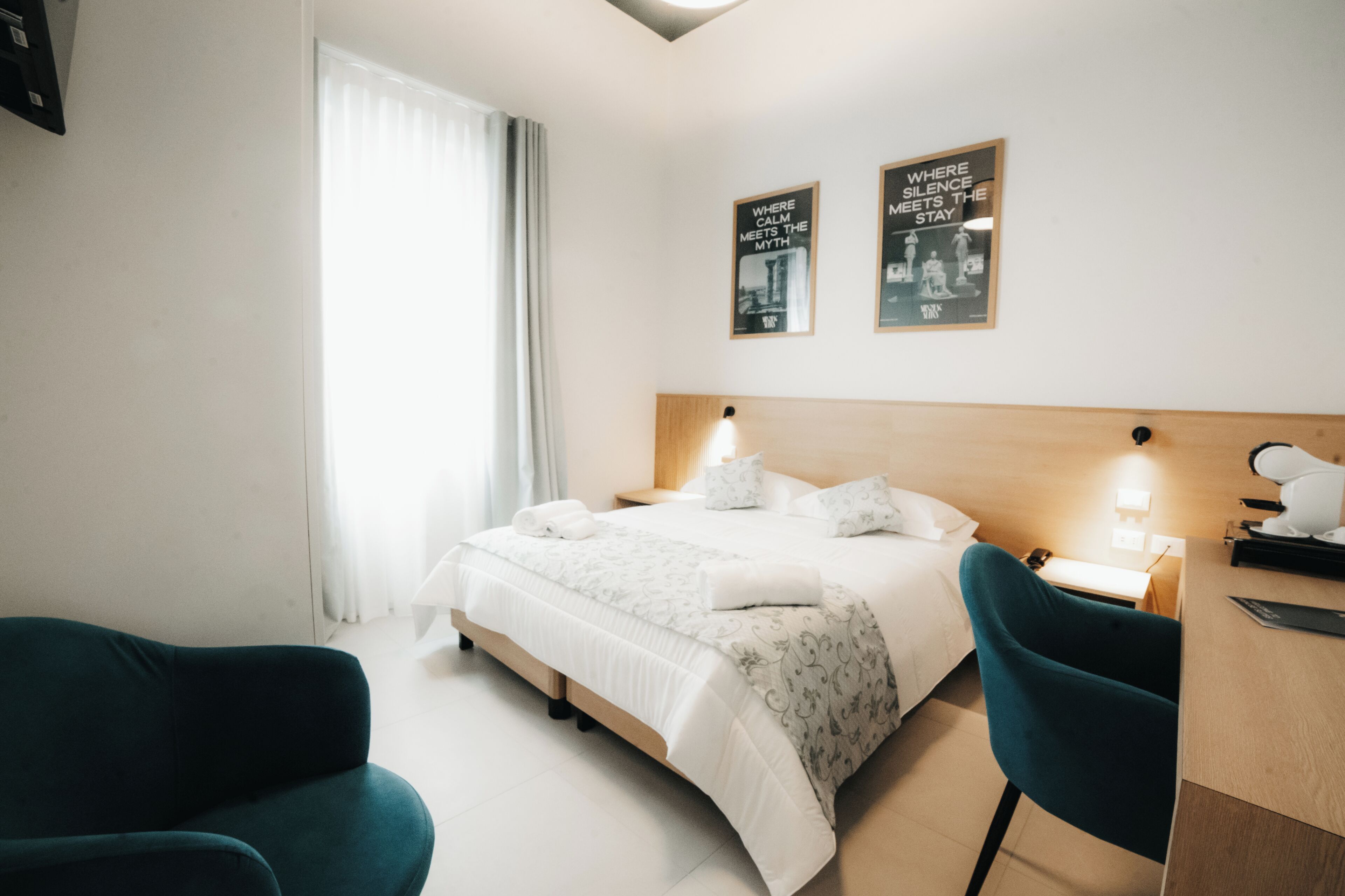 A hotel room with a bed, two wall-mounted lights, a desk, a chair, and two posters on the wall.