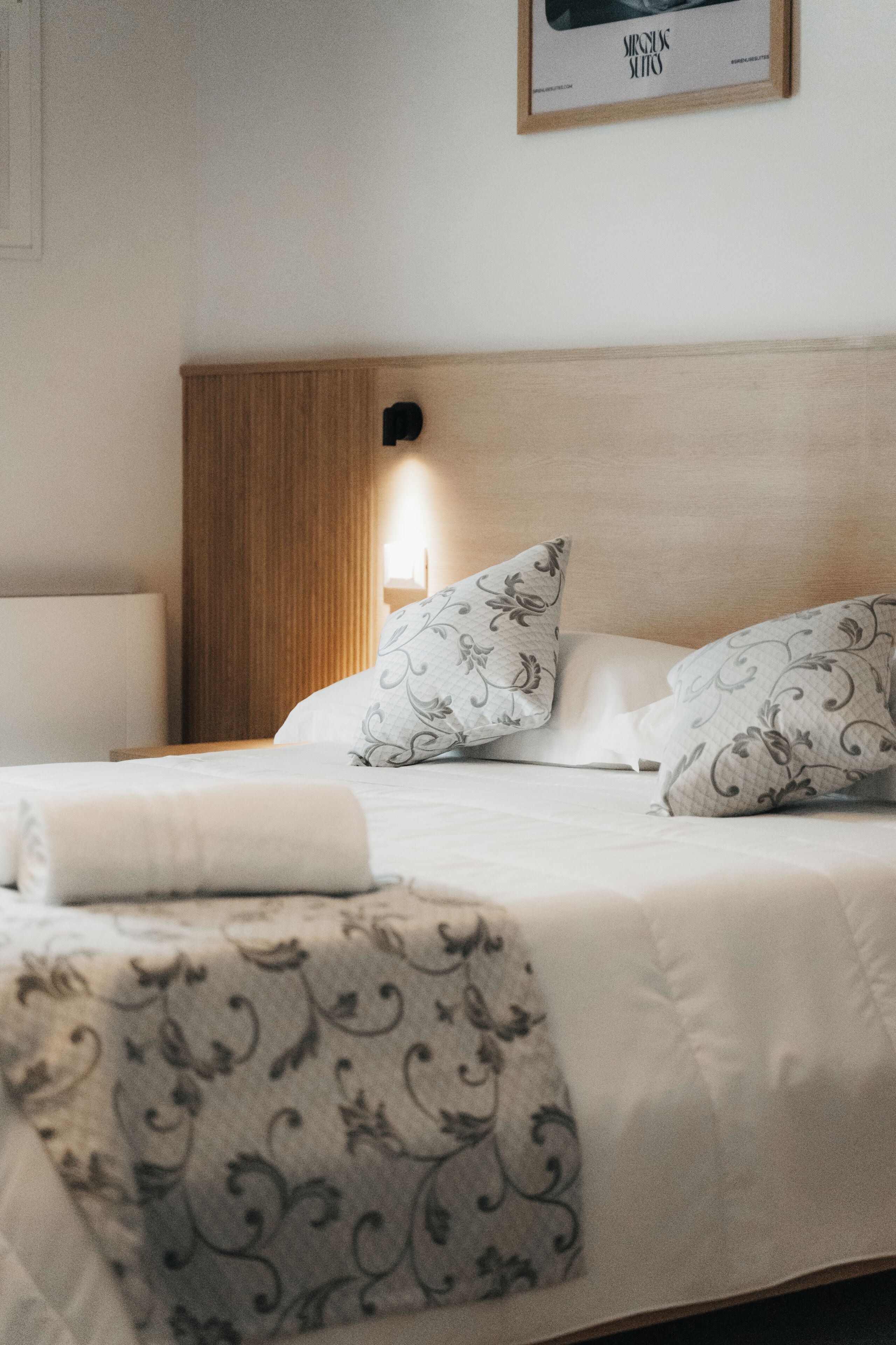 Una cama bien hecha con almohadones y una manta estampada, un cabecero de madera y una lámpara fijada a la pared.